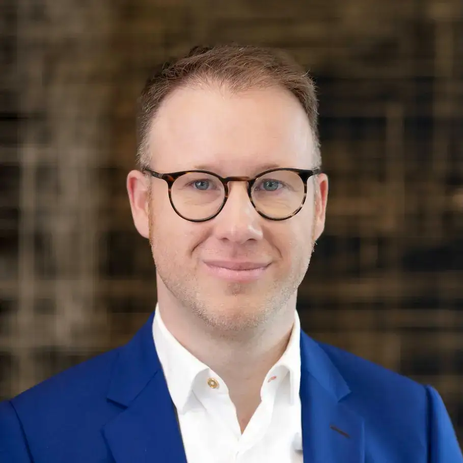Mann in Brille und blauer Jacke vor unscharfem Hintergrund.