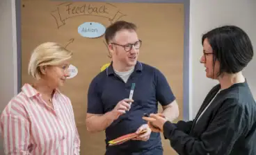 Drei Menschen im Gespräch vor einer Tafel mit Feedback-Plan.