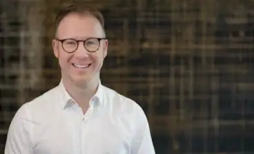 Lächelnder Mann mit Brille vor dunklem Hintergrund.