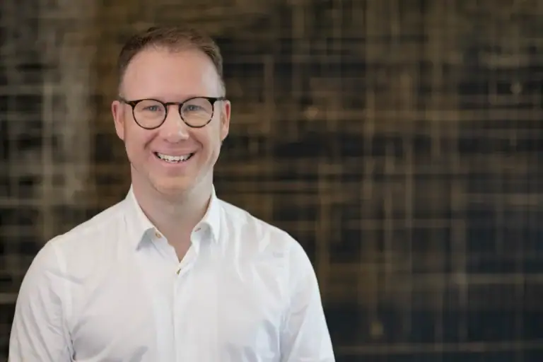 Lächelnder Mann mit Brille vor dunklem Hintergrund.