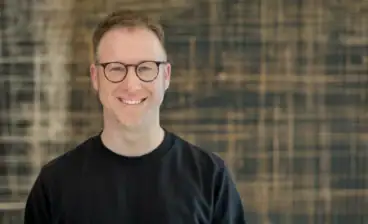 Mann mit Brille und schwarzem Pullover vor Holz-Hintergrund.